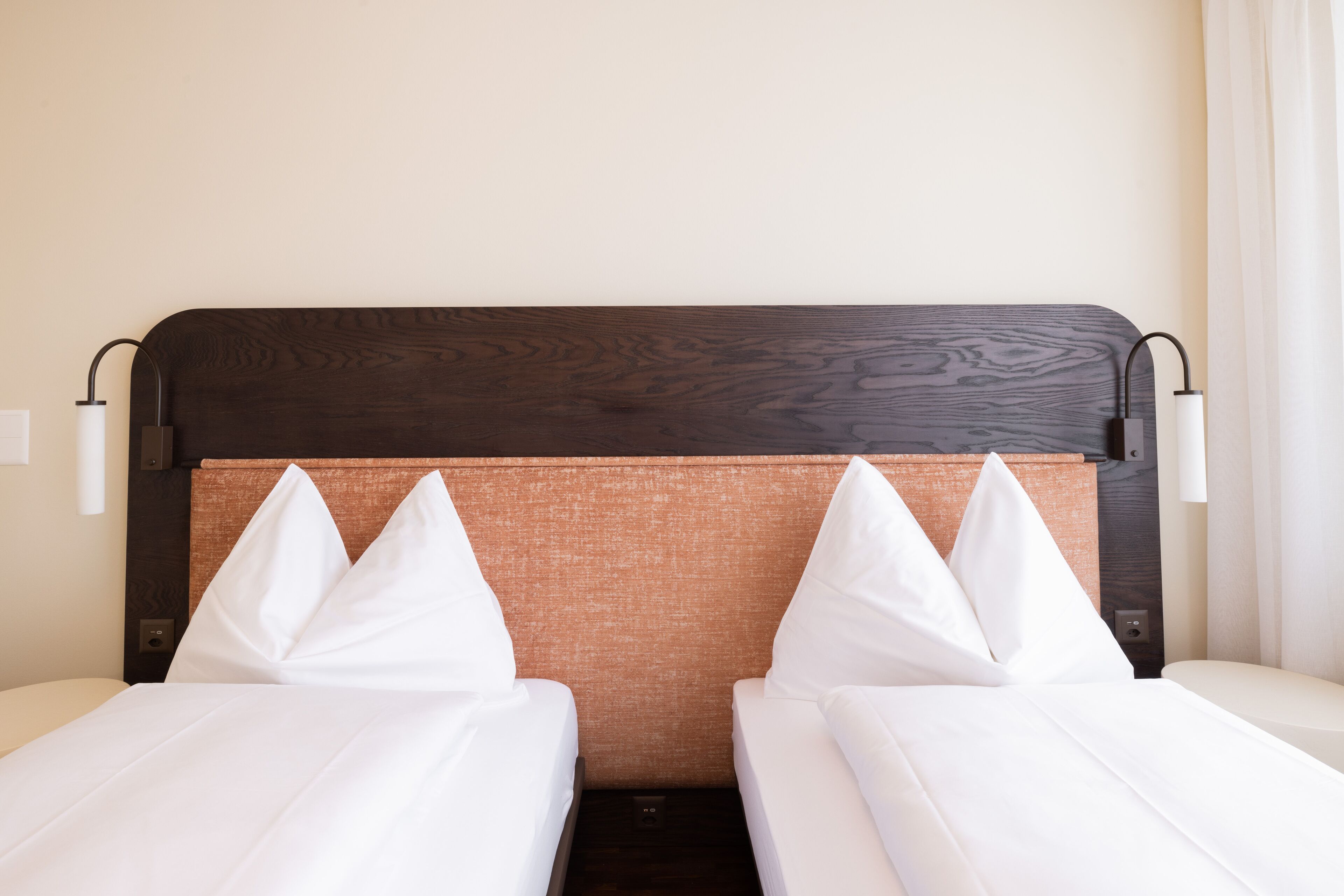 Une chambre d’hôtel avec deux lits simples, une tête de lit en bois et une tête de lit recouverte d’un tissu marron.