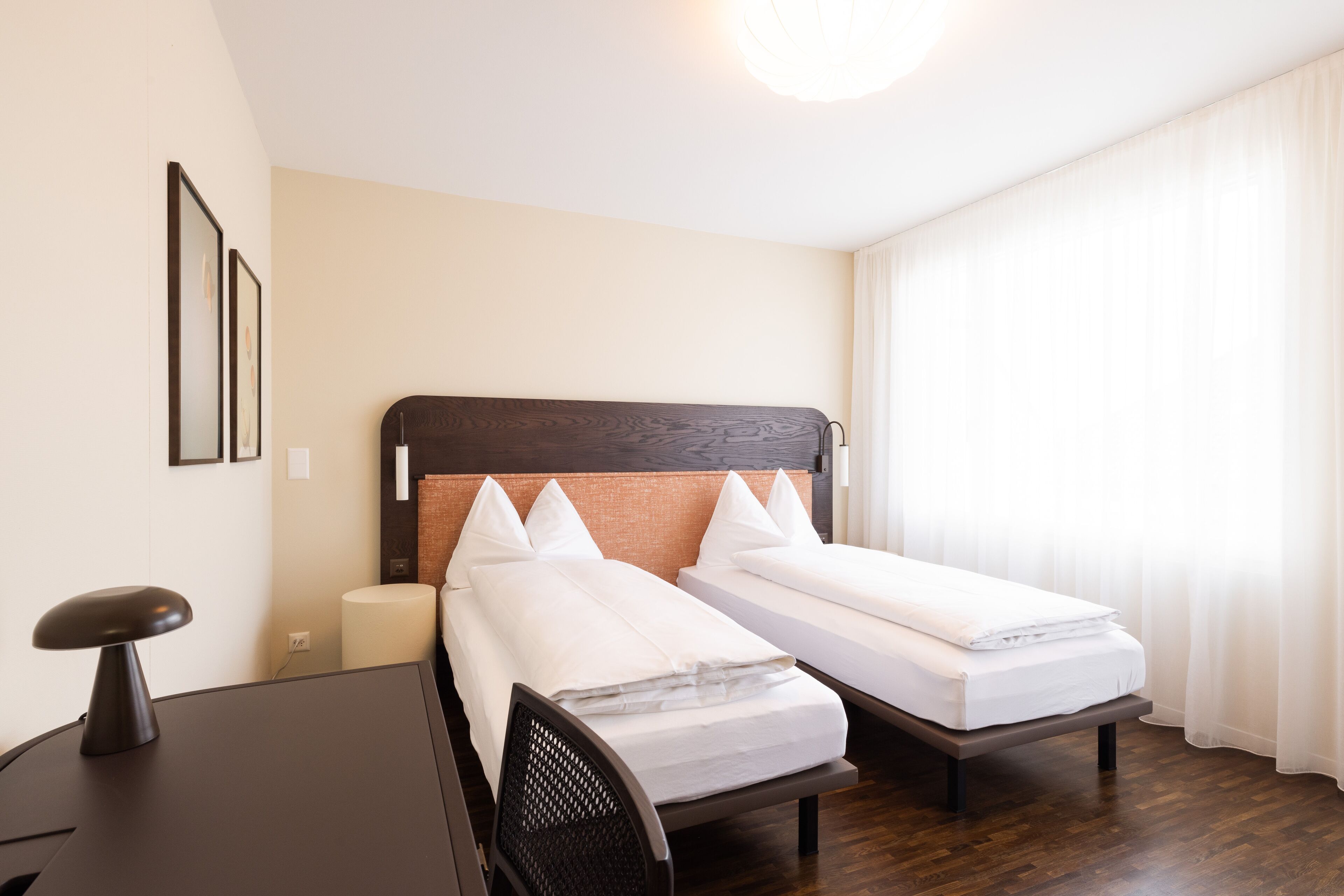 A hotel room with two single beds, a desk, a chair, a lamp, and a large window with curtains.