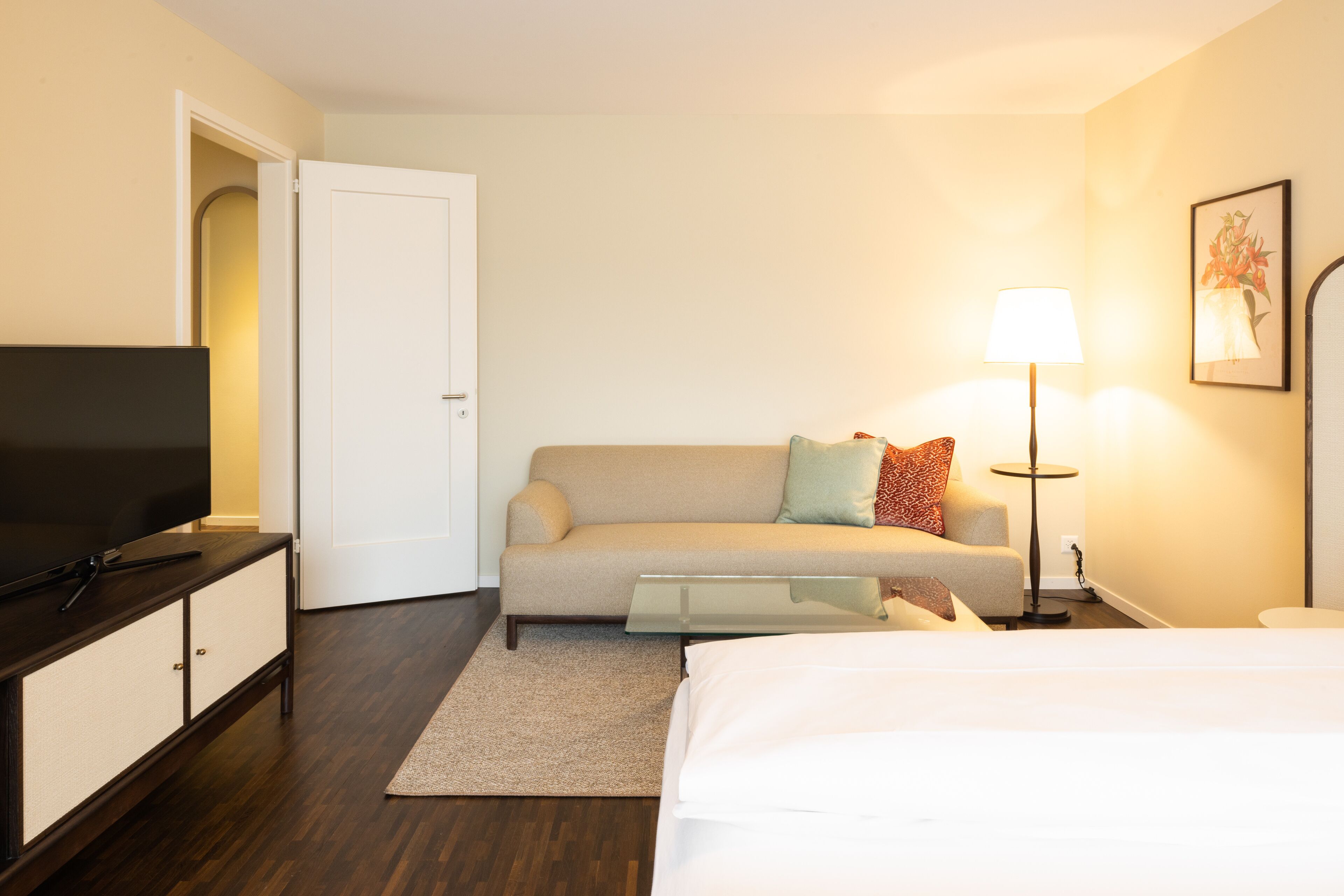 A hotel room with a bed, a sofa, a TV on a cabinet, a lamp, and a painting on the wall.