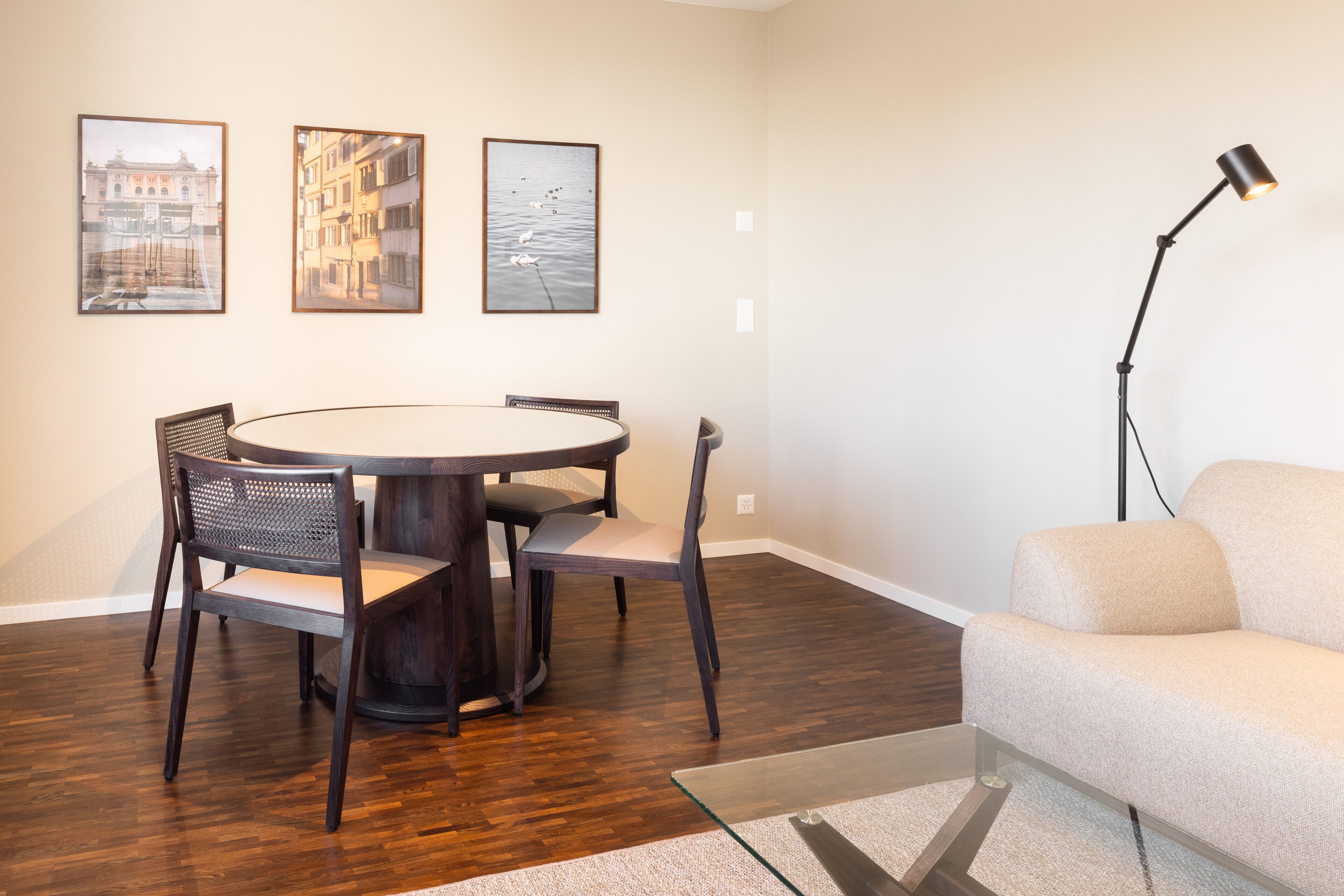 A room with a round dining table, four chairs, a sofa, and three framed pictures on the wall.