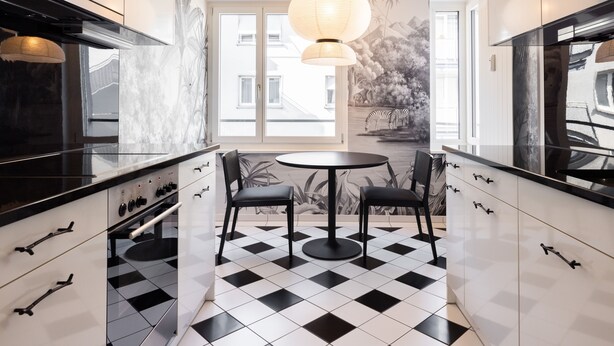 Een moderne keuken met zwart-wit geruit vloerkleed, twee zwarte stoelen rond een klein tafeltje, en een groot raam met uitzicht op gebouwen en bomen.