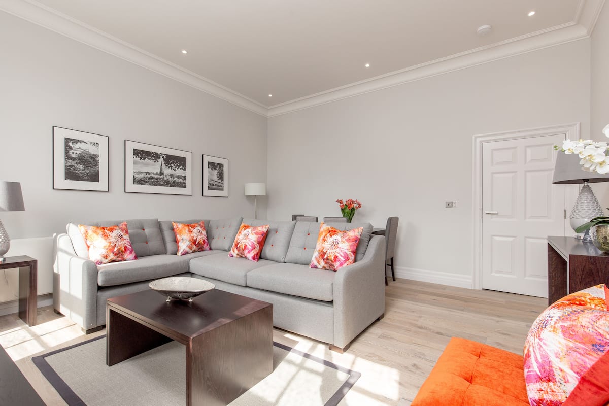 A modern living room with a grey sectional sofa, a coffee table, and a vibrant orange ottoman. There are three framed photographs on the wall, a white door, and a small side table with a lamp.