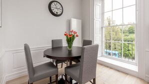 A dining area with a round table, four chairs, a vase with flowers, a clock, and a large window with a view of greenery and a blue vehicle. - Destiny Scotland Chisholm Hunter Suites (Edinburgh)