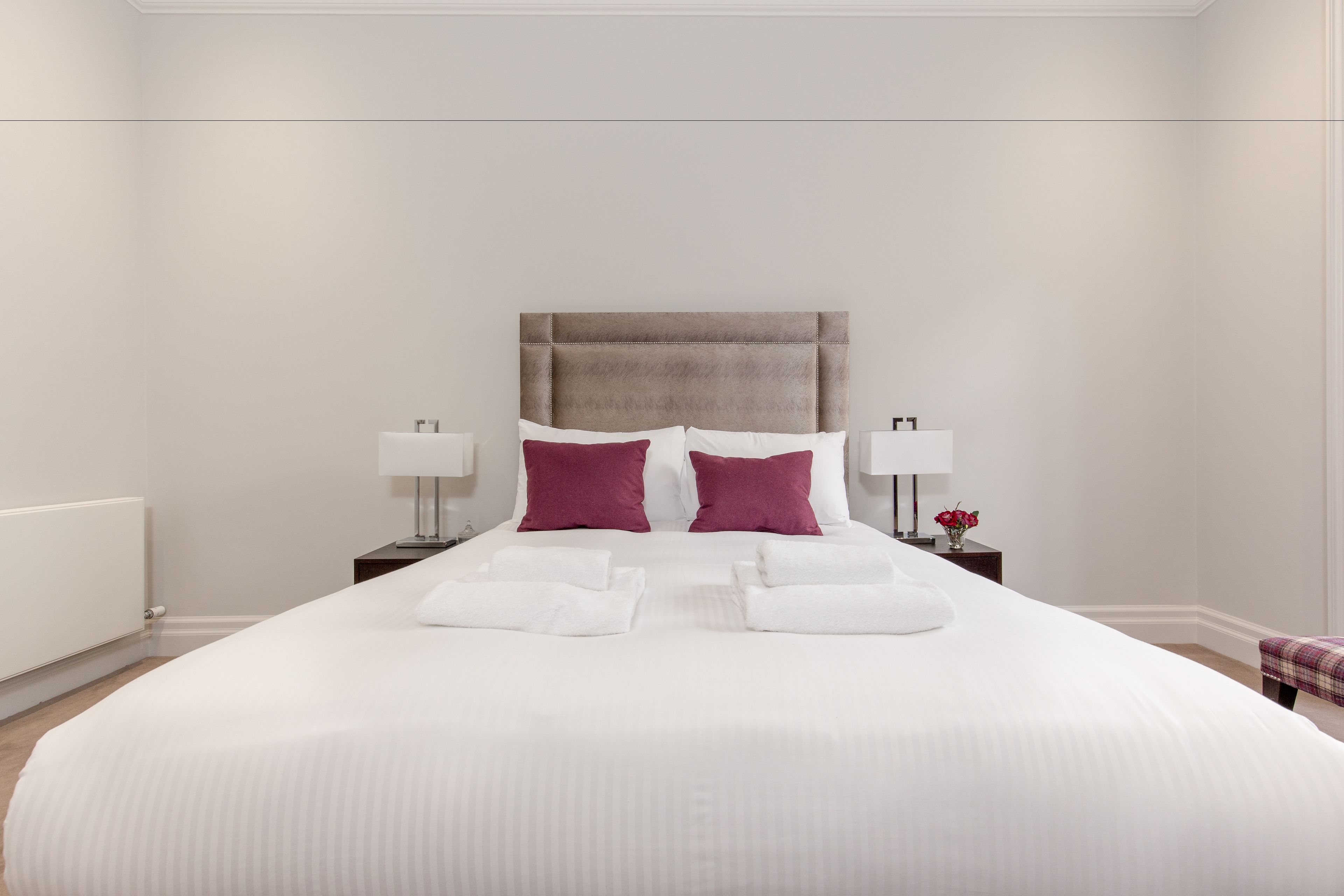 A neatly made bed with white bedding and two pillows, a bedside table with a lamp, and a small vase with flowers.