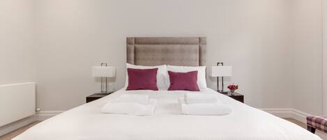 A neatly made bed with white bedding and two pillows, a bedside table with a lamp, and a small vase with flowers.