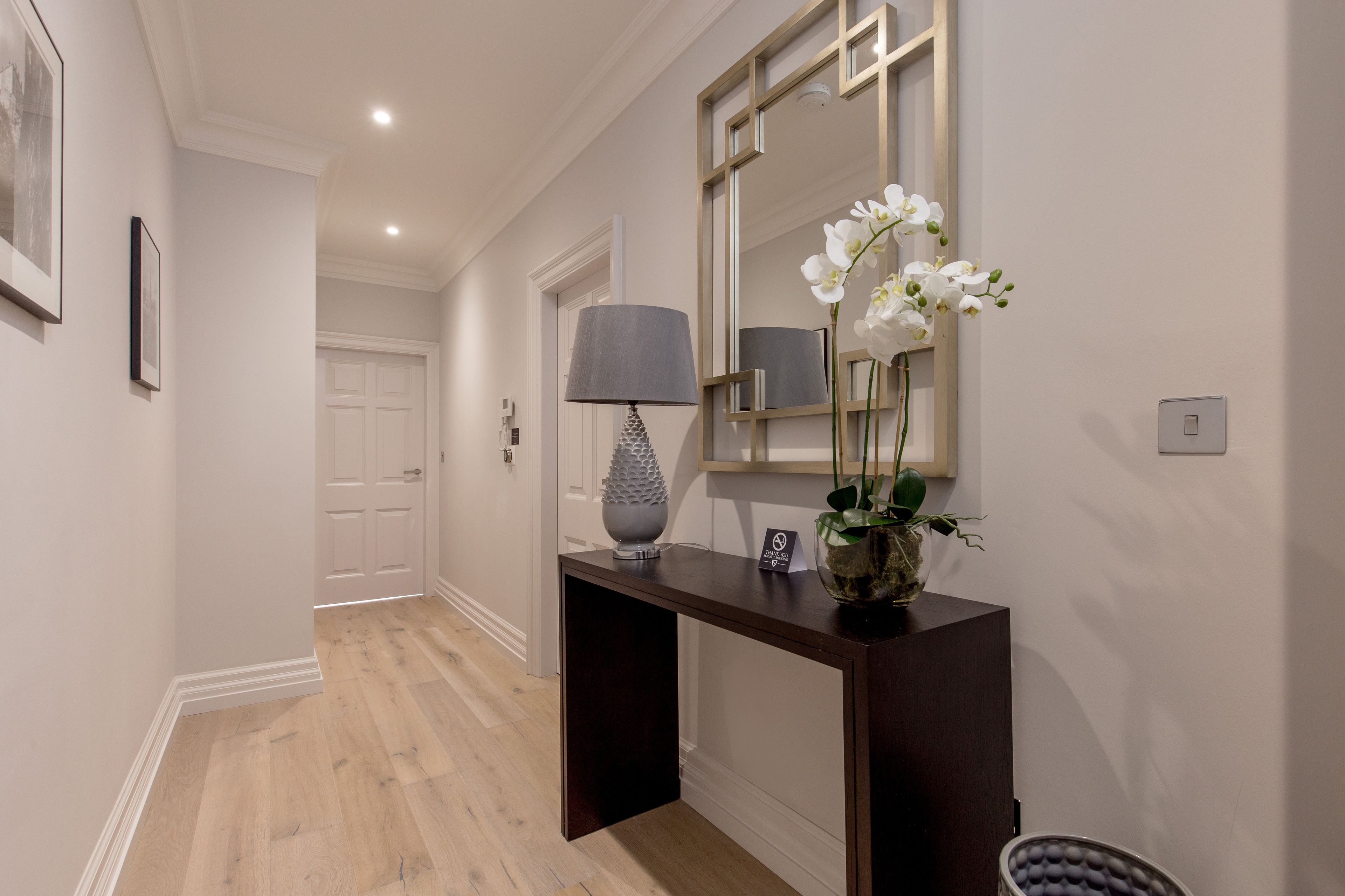 A hallway with a wooden floor, a dark console table with a lamp, a mirror, and a vase with white flowers.