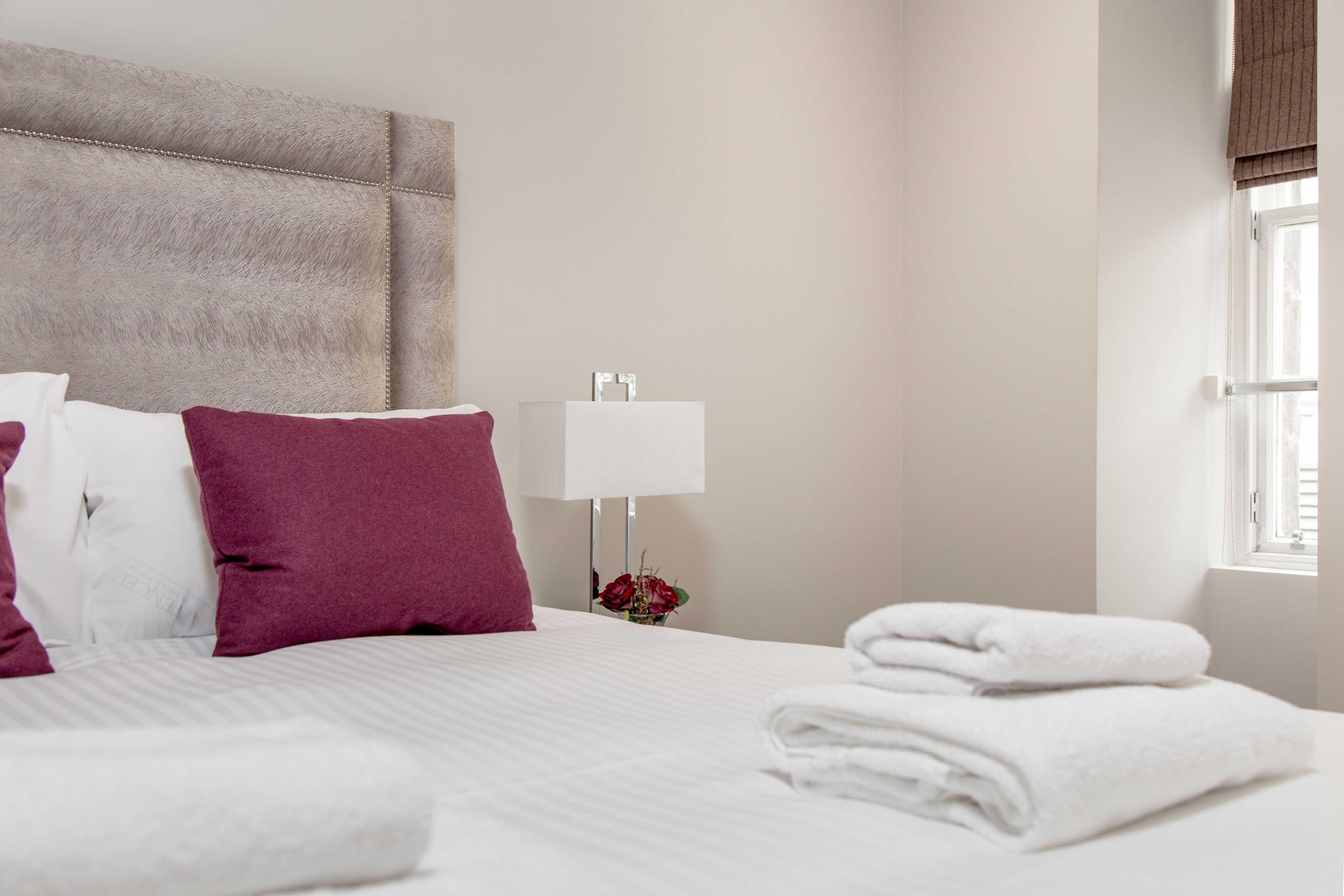 Uma cama bem arrumada com almofadas brancas e bordô, um abajur branco e uma janela com persianas.