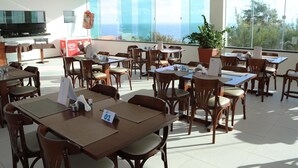 Un amplio comedor con múltiples mesas y sillas, ventanas grandes con vistas al océano y una zona de cocina con televisor de pantalla plana.