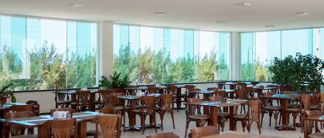 A spacious indoor area with multiple wooden tables and chairs arranged in rows, large windows providing natural light and views of greenery outside.