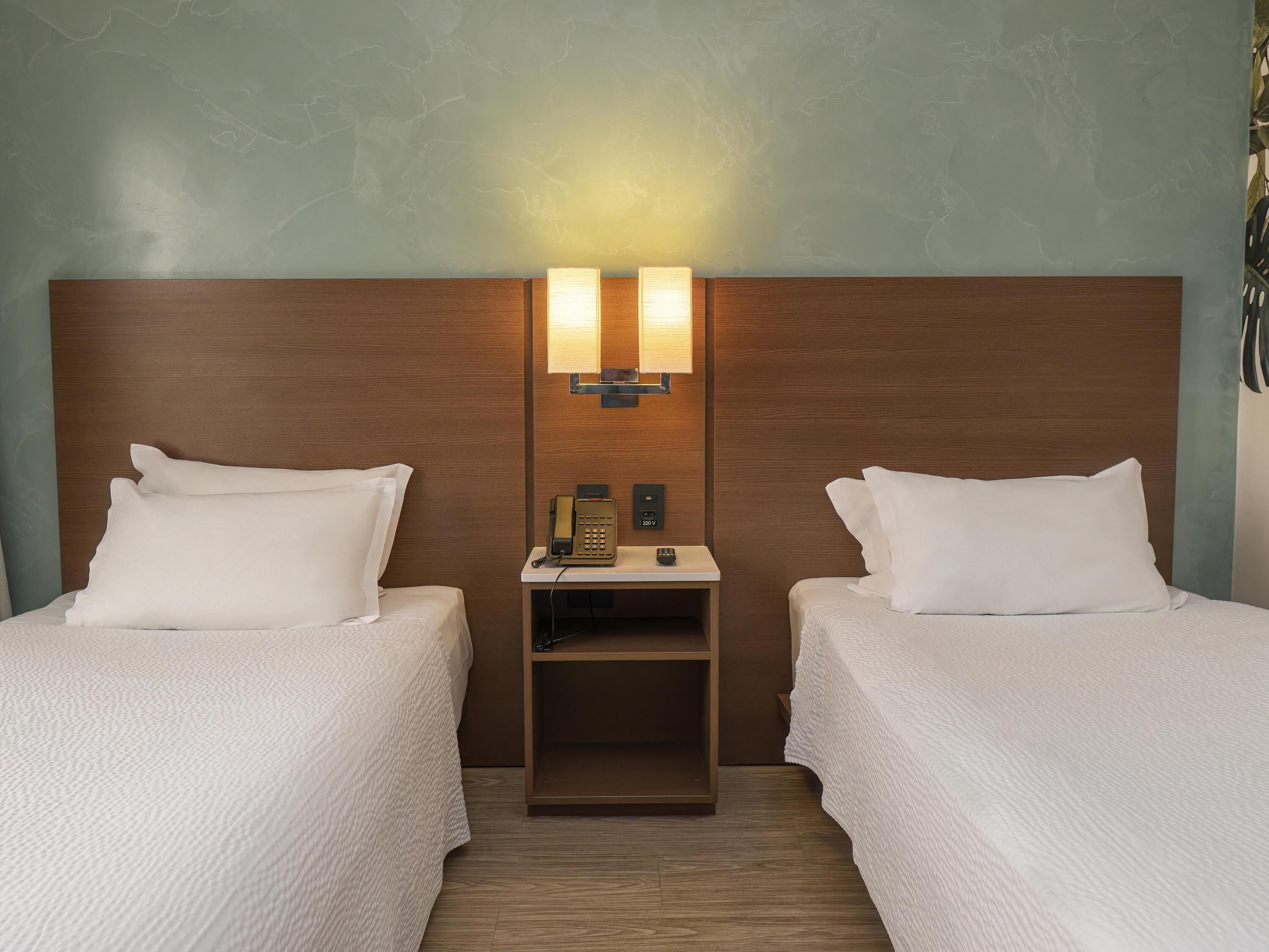A hotel room with two beds, a nightstand with a phone, and a wall-mounted light fixture.