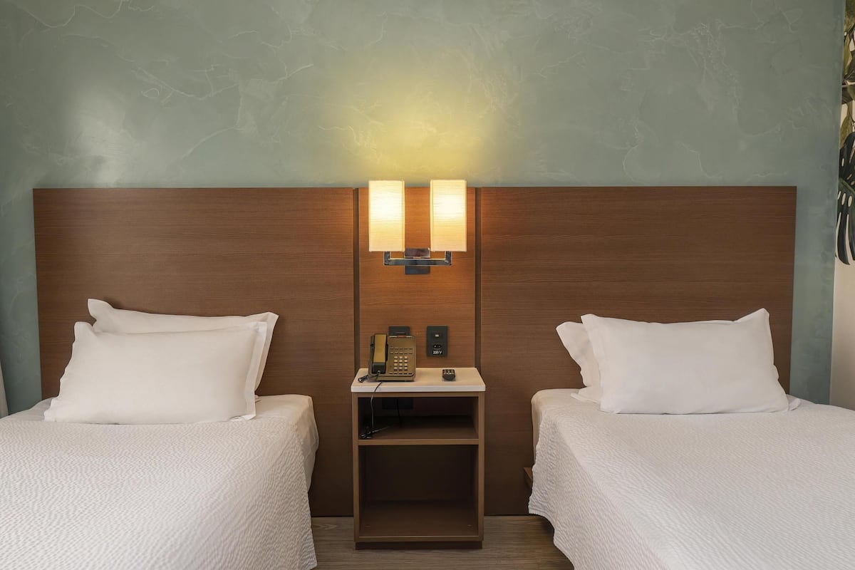 A hotel room with two beds, a nightstand with a phone, and a wall-mounted light fixture.