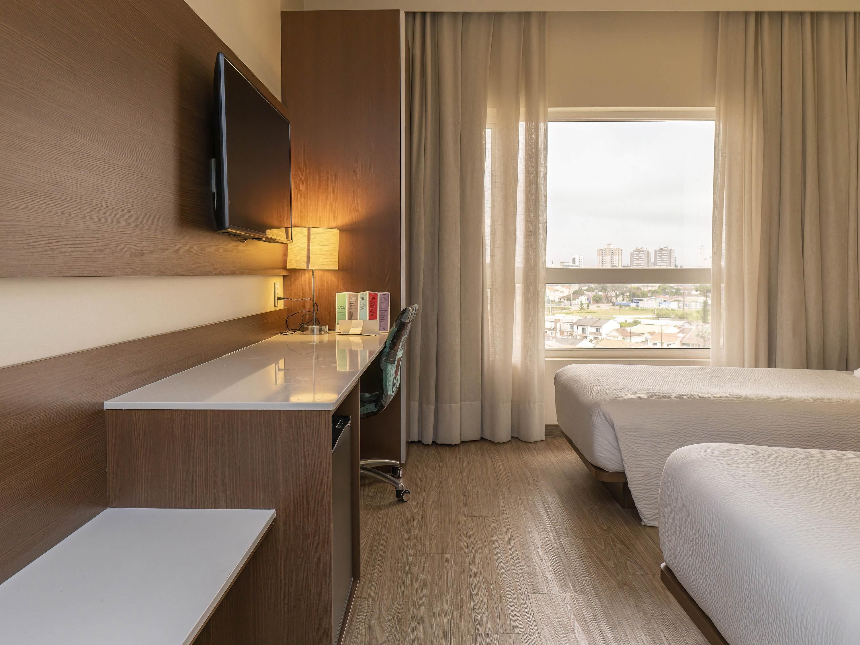 A hotel room with a desk, a flat-screen TV, a lamp, a bed, and a window with curtains.
