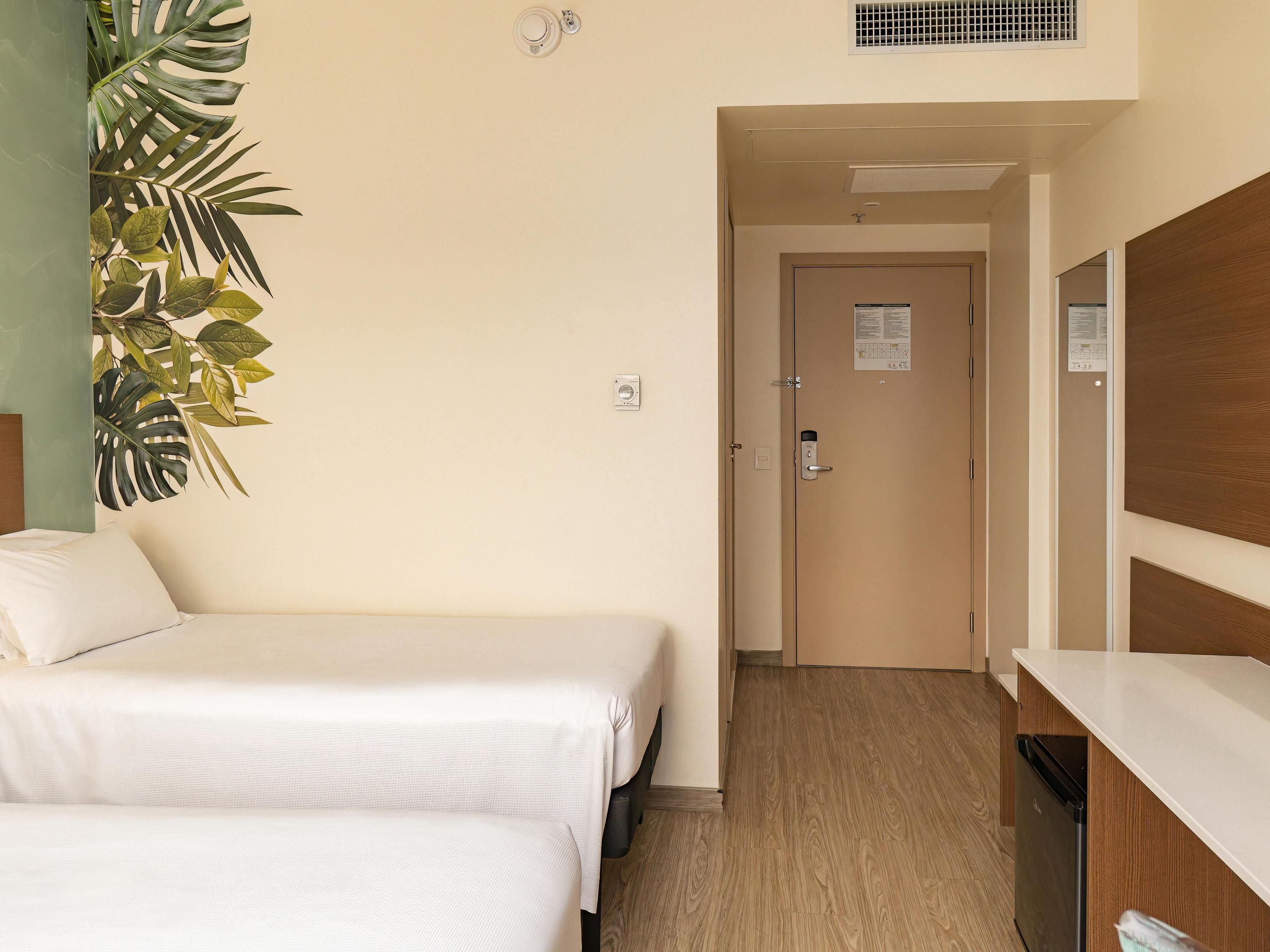 Habitación de hotel con dos camas, un mostrador de madera, una puerta con cerradura y una planta en la pared.