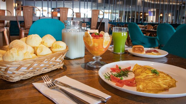 A dining table with a basket of bread rolls, a glass of milk, a fruit drink, and a plate with an omelette and vegetables. - ibis Styles Curitiba Aeroporto (Sao Jose dos Pinhais)