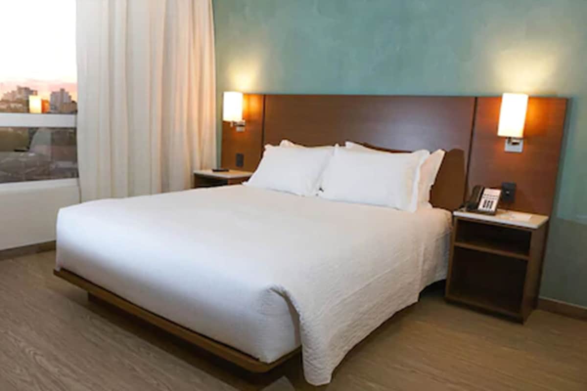 Een hotelkamer met een groot bed, twee nachtkastjes, een raam met gordijnen en uitzicht op de stad.