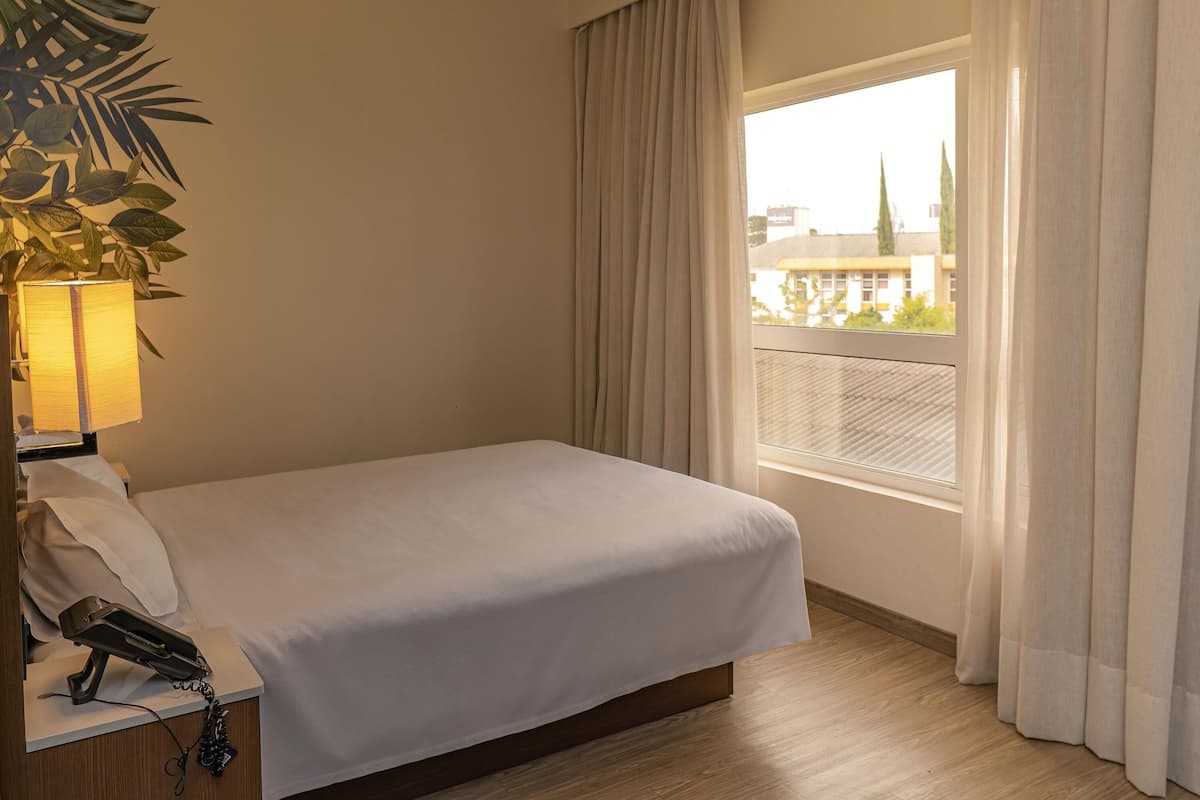 Une chambre d’hôtel avec un lit, une lampe de chevet, un téléphone et une fenêtre avec des rideaux.