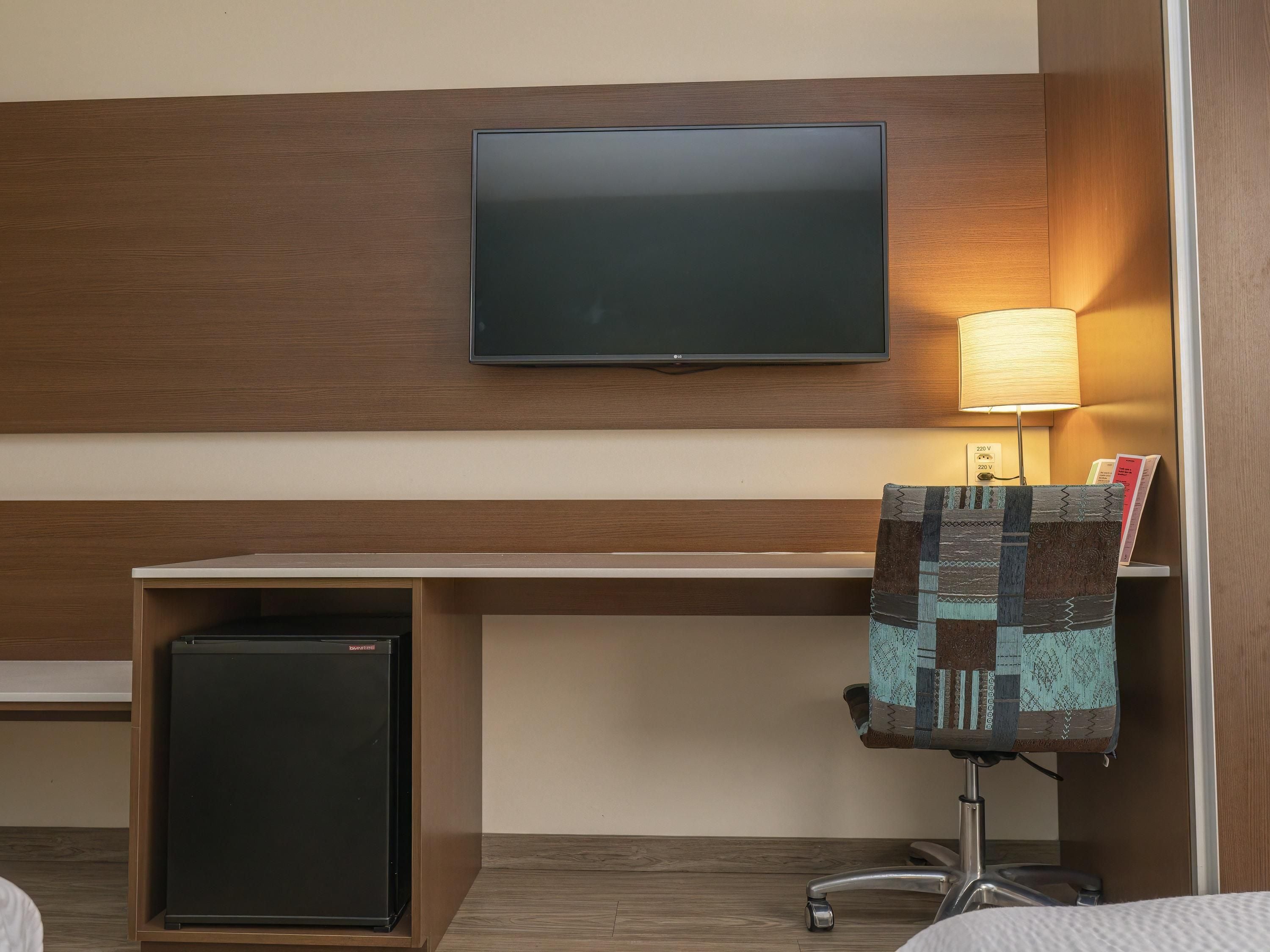 Een hotelkamer met een wandgemonteerde flatscreen-tv, een bureau met lamp, een kleine koelkast en een gestreept fauteuil.