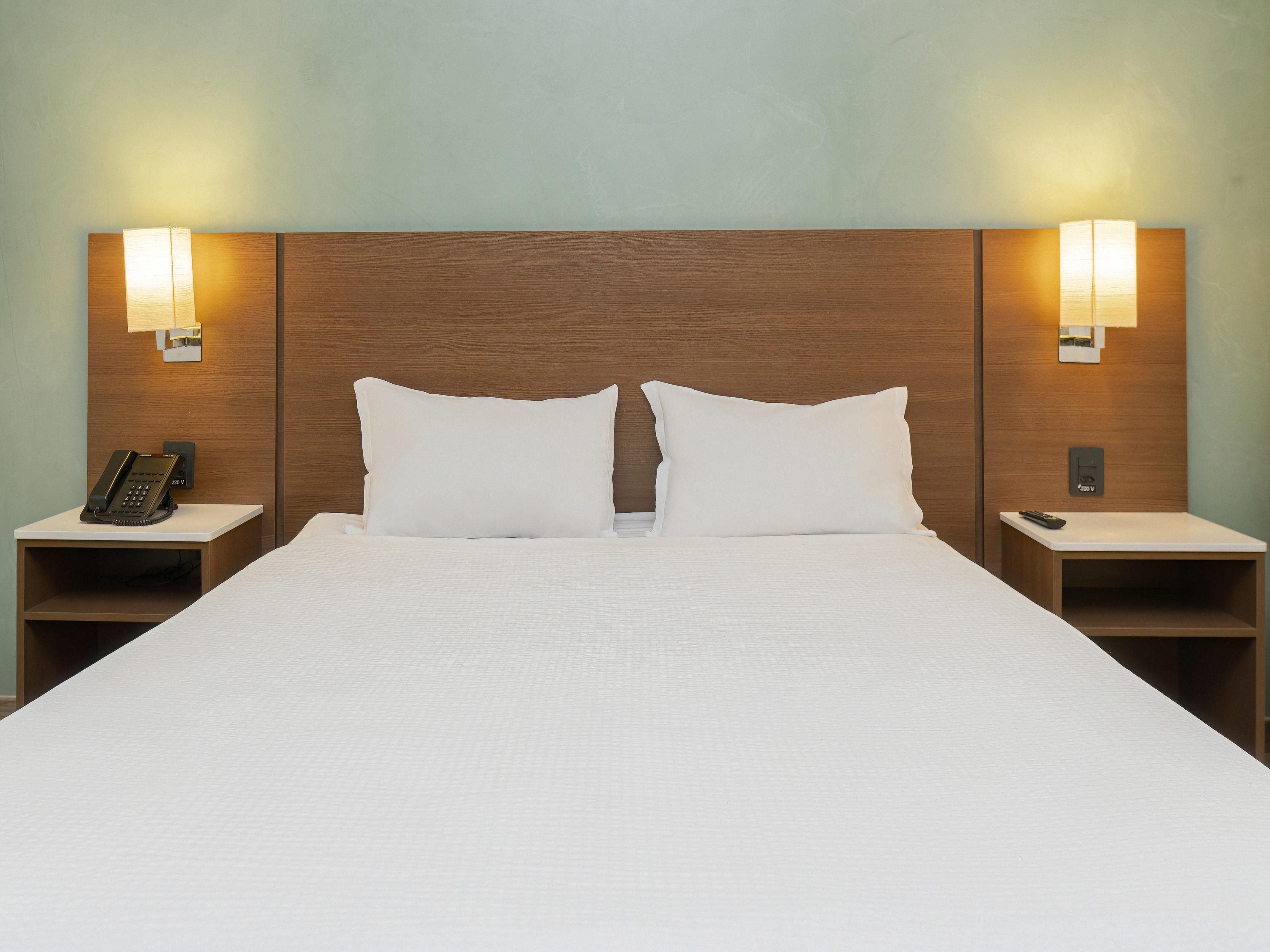 Een hotelkamer met een groot bed, twee nachtkastjes, een telefoon op het linker nachtkastje en een afstandsbediening op het rechter nachtkastje.