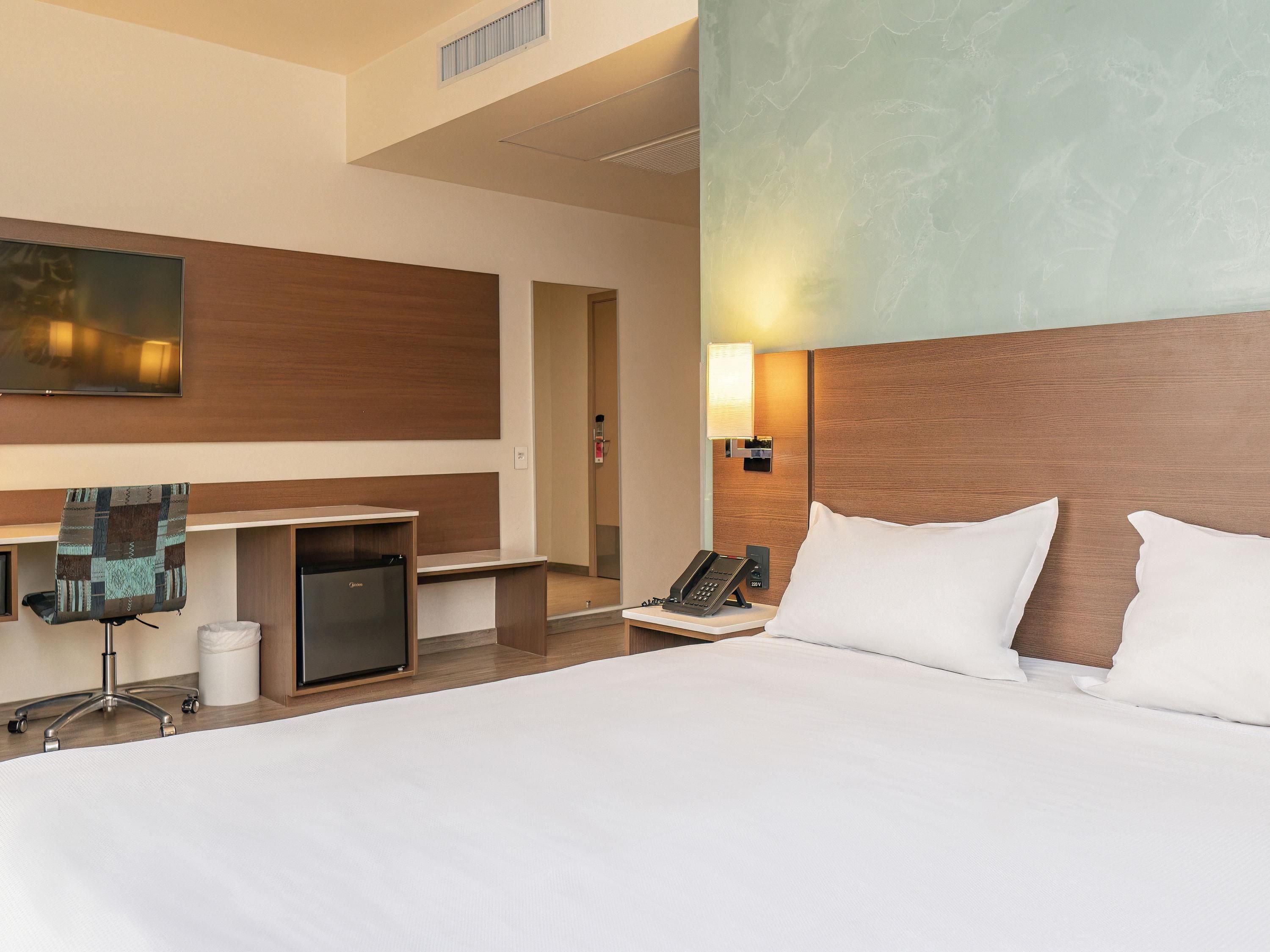 A hotel room with a large bed, a desk with a chair, a telephone, and a wall-mounted TV.