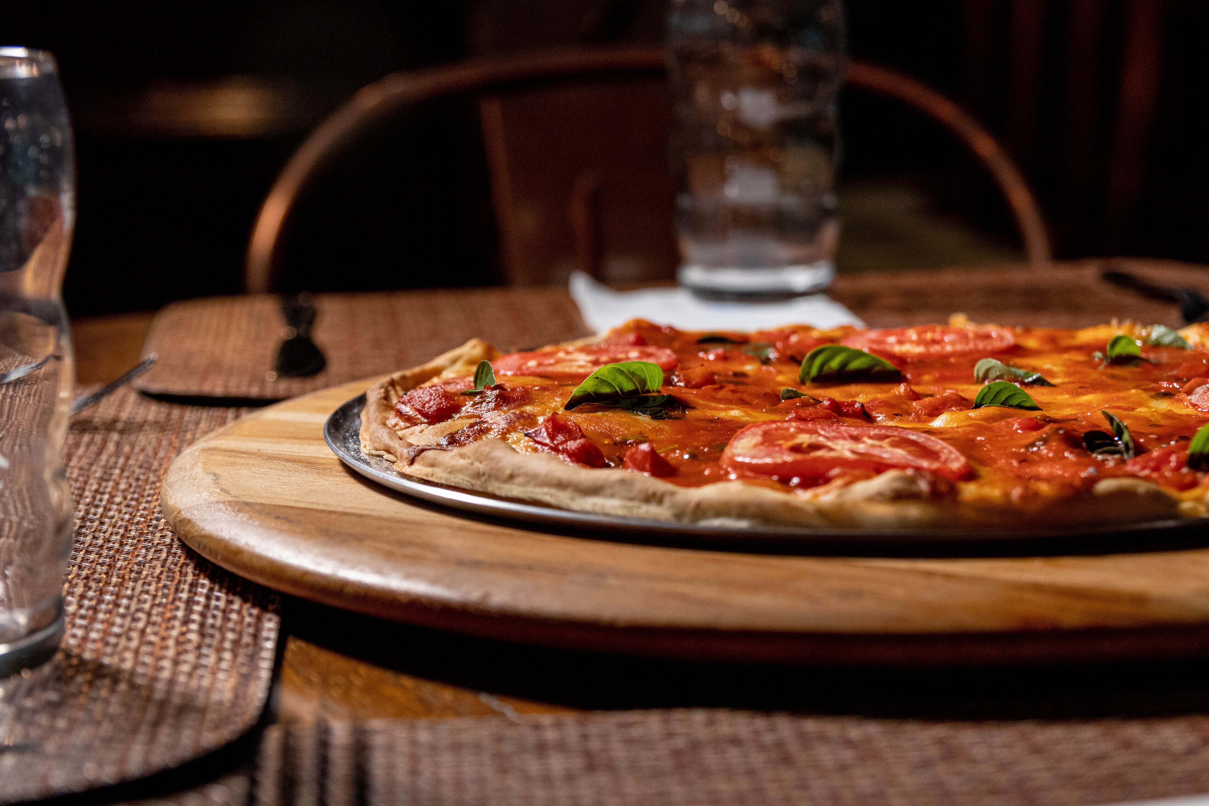 A pizza on a wooden board on a table with a glass of water and a chair in the background.