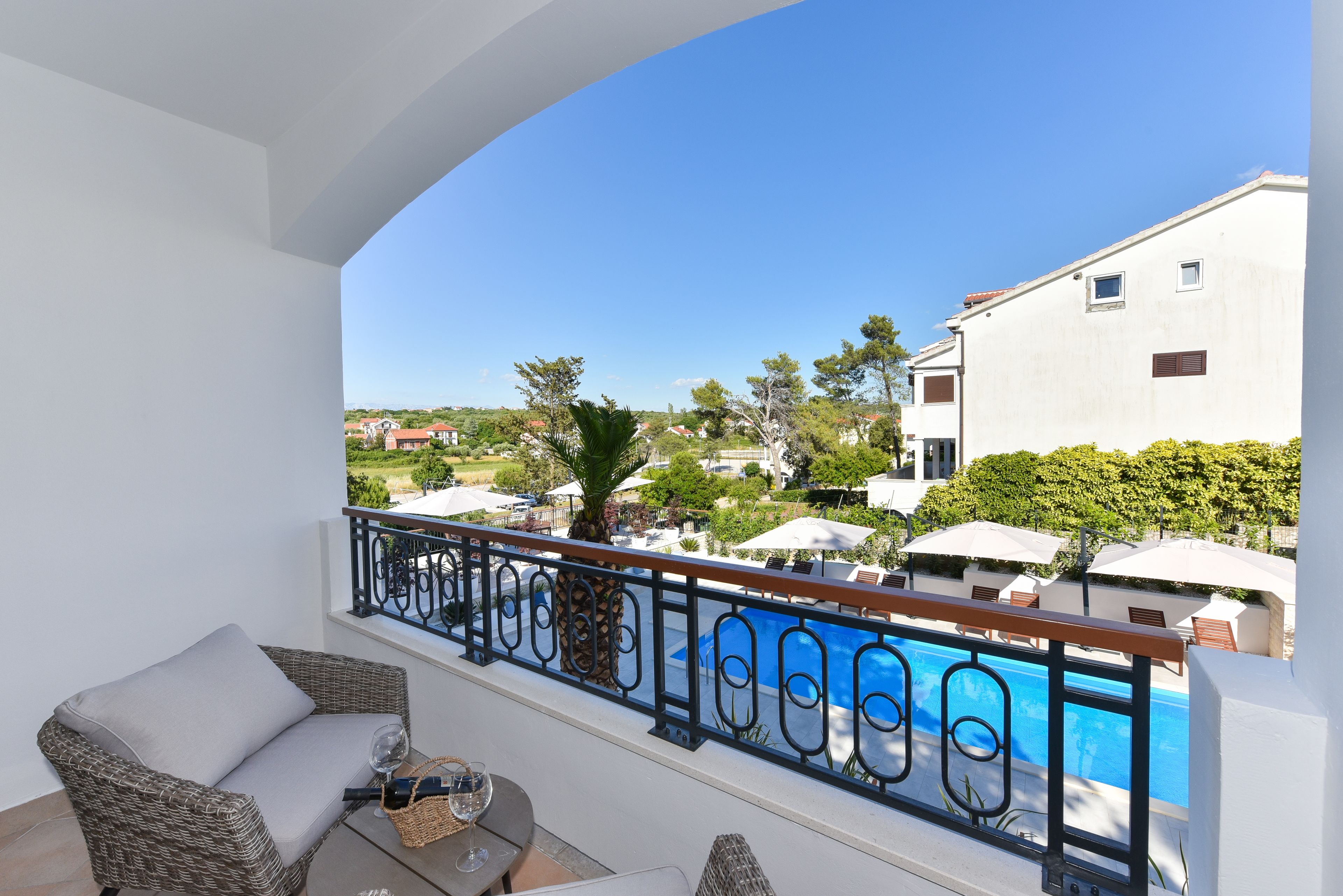 A balcony with wicker furniture, a glass table, and a view of a pool and buildings.