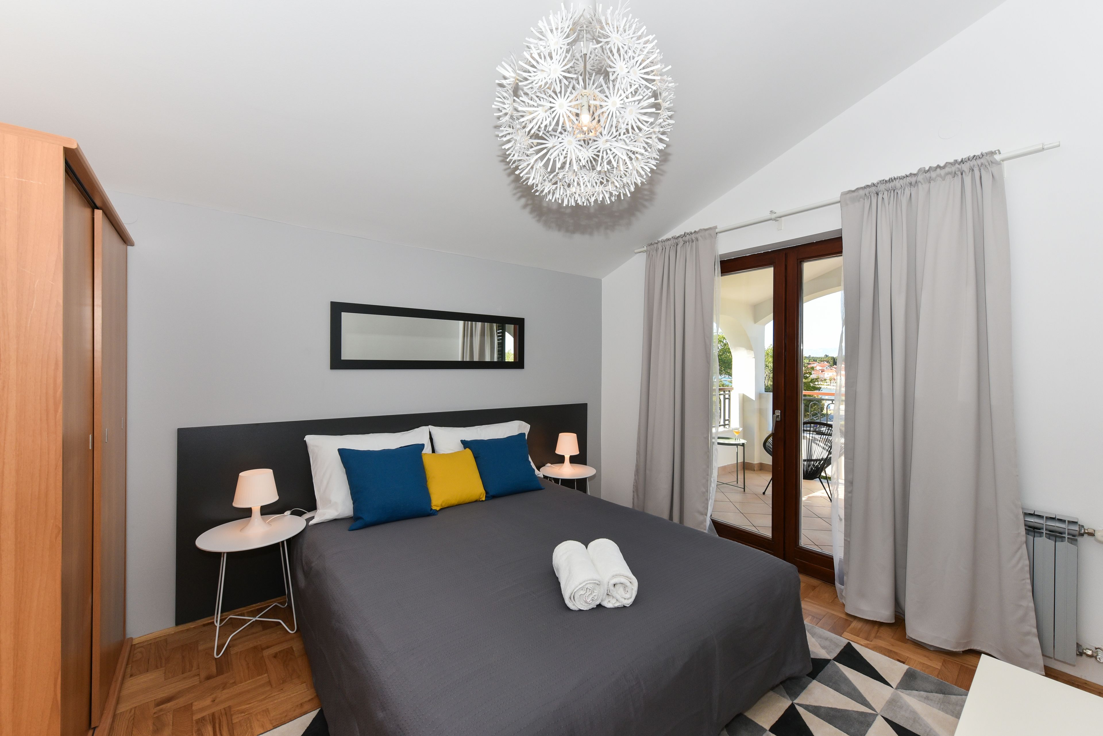 A modern bedroom with a large bed, a bedside table with a lamp, a mirror on the wall, and a view of the outside through a sliding door.
