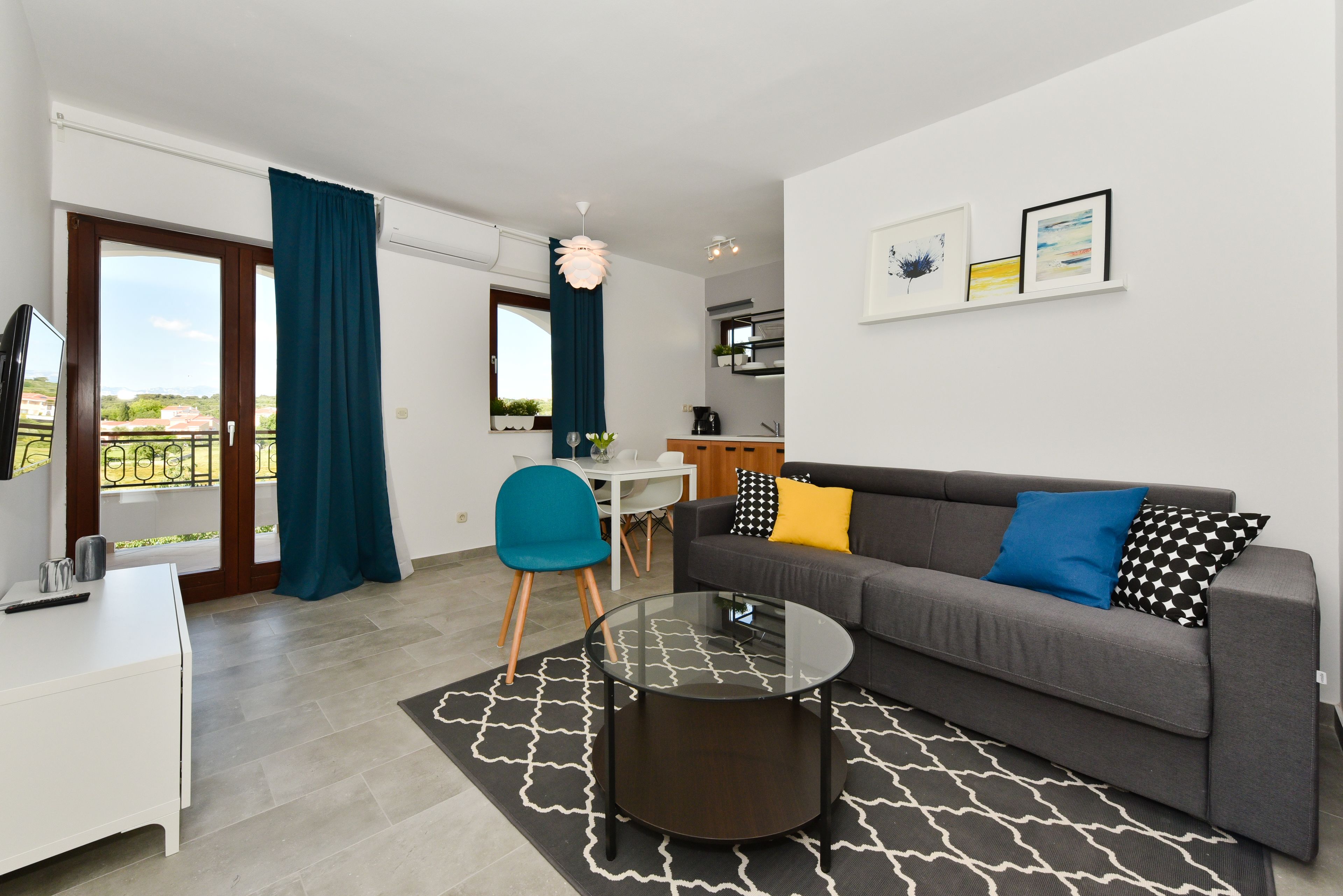 Ein modernes Wohnzimmer mit grauem Sofa, einem Glastisch, einem weißen Schreibtisch und einem blauen Sessel. Ein Balkon bietet Blick auf die Stadt.