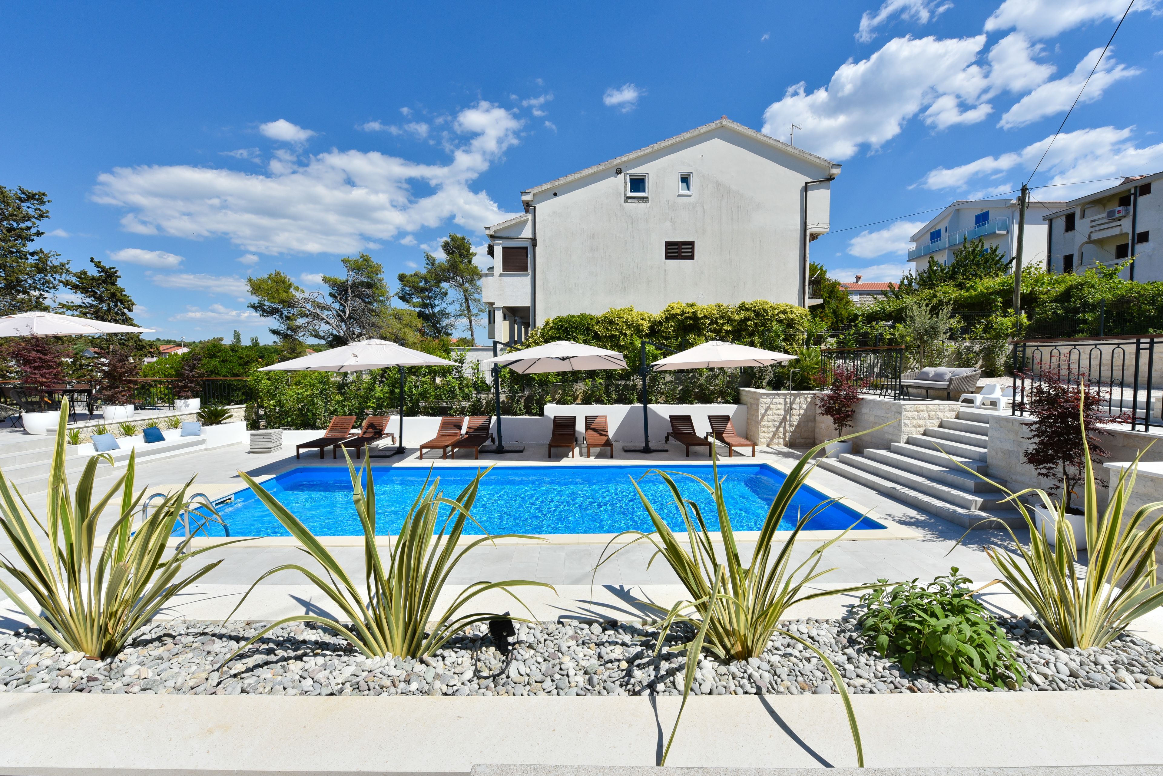 A swimming pool with lounge chairs and umbrellas, surrounded by plants and a building in the background.