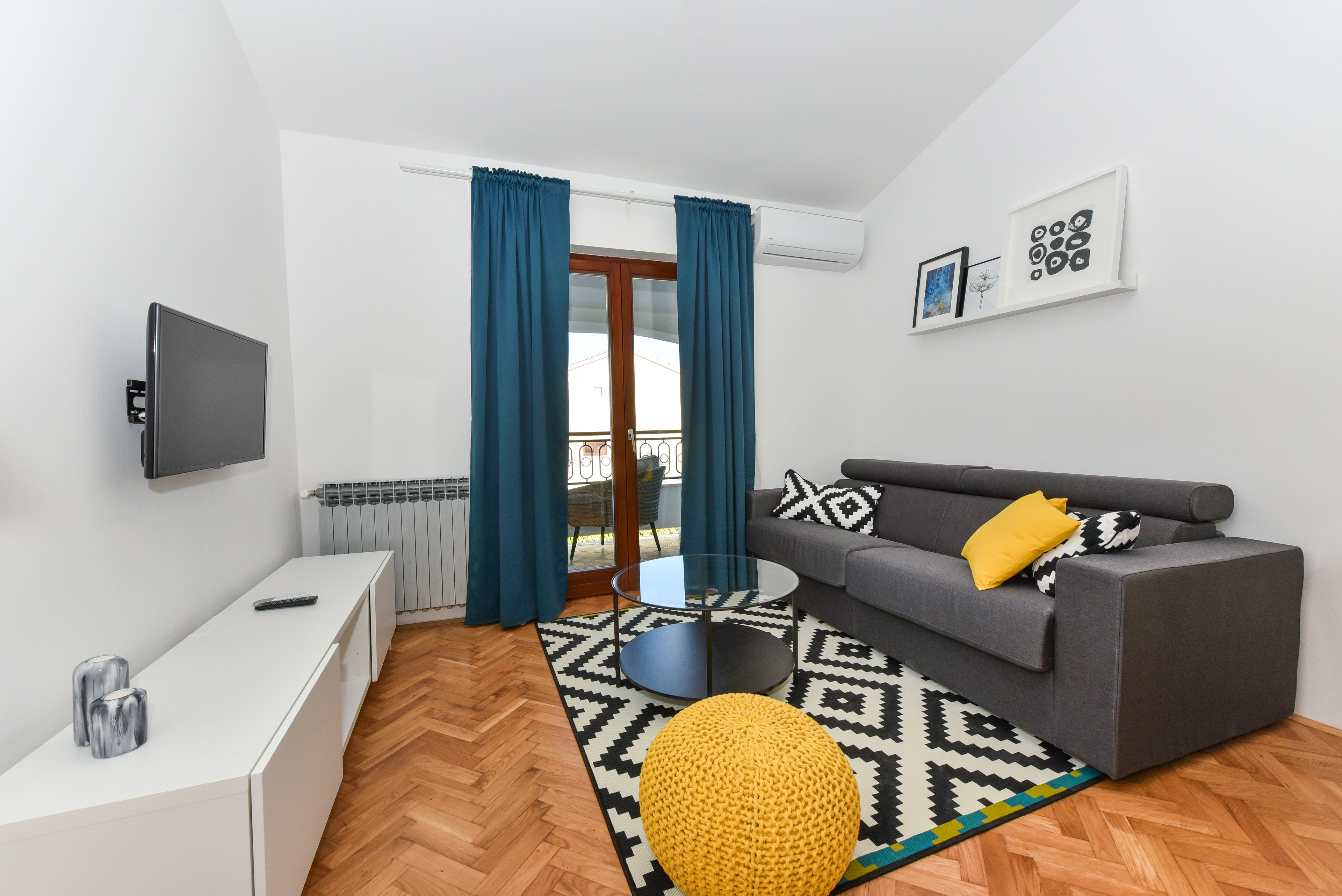 Ein modernes Wohnzimmer mit einem grauen Sofa, einem runden Couchtisch, einer weißen TV-Konsole und einem gemusterten Teppich.