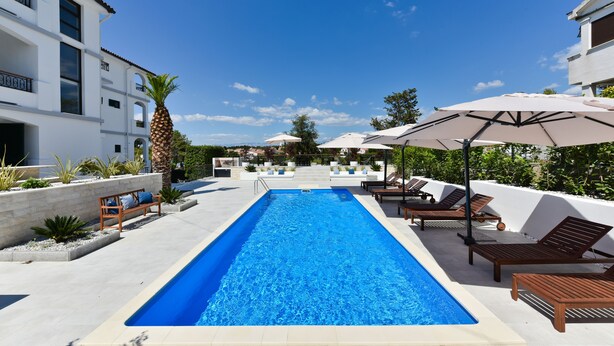 A rectangular swimming pool with lounge chairs and umbrellas on one side, and a building with balconies on the other. - Mediterraneo Garden Apartments (Zadar)