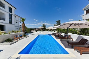 A rectangular swimming pool with lounge chairs and umbrellas on one side, and a building with balconies on the other. - Mediterraneo Garden Apartments (Zadar)