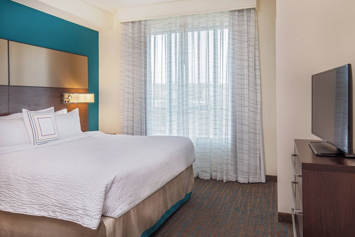 A hotel room with a large bed, a television on a wooden cabinet, and a window with sheer curtains.