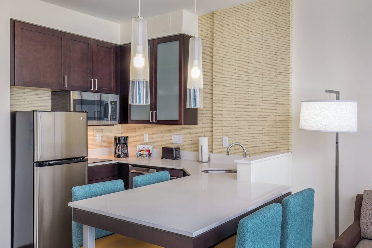 A modern kitchen with dark wooden cabinets, stainless steel appliances, a central island with blue chairs, and a tall floor lamp.