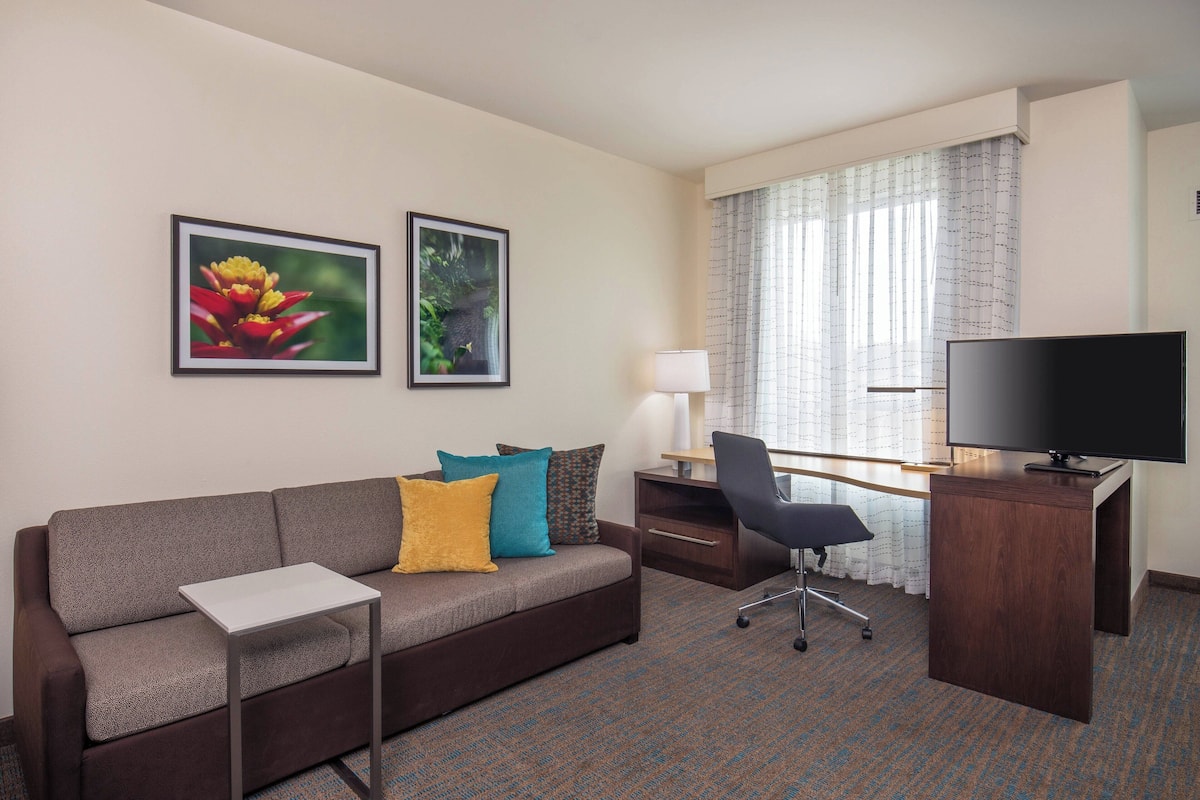 A hotel room with a sofa, a desk with a TV, a lamp, and two framed pictures on the wall.