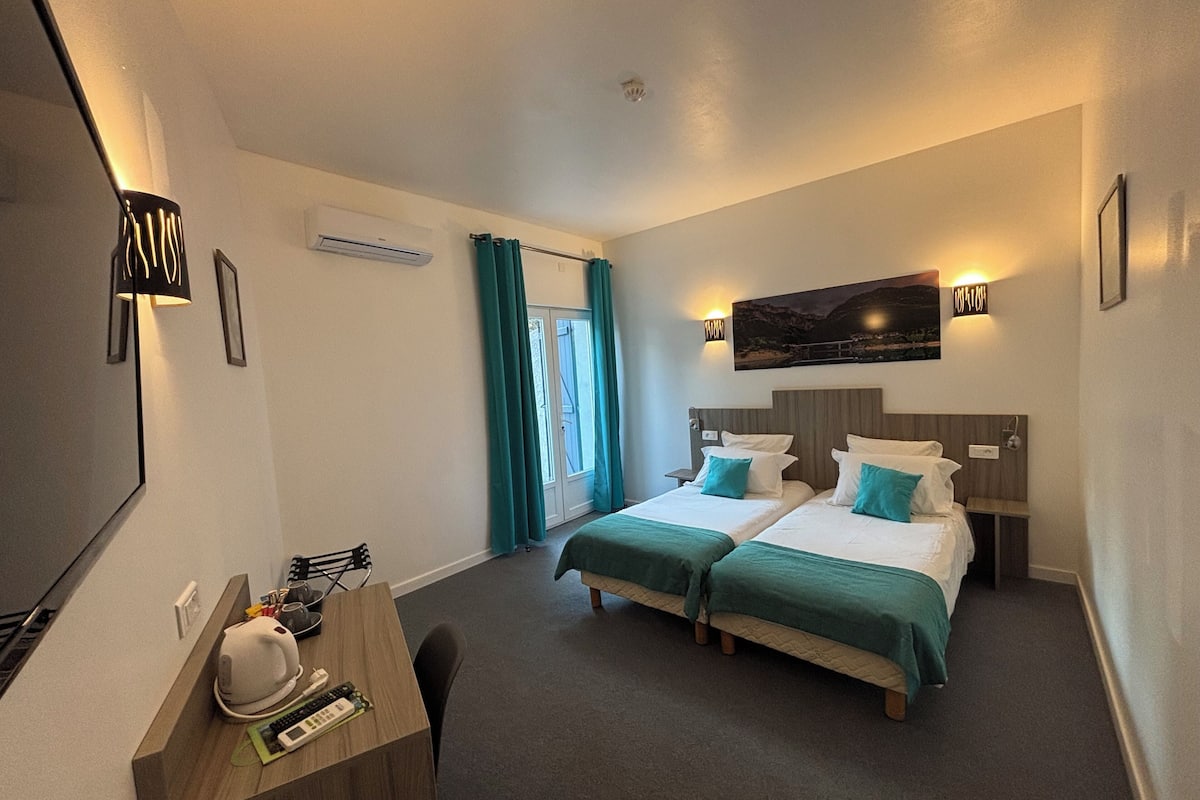 Ein Hotelzimmer mit zwei Betten, einem Schreibtisch mit Stuhl, einem Fernseher und einem Fenster mit Vorhängen.