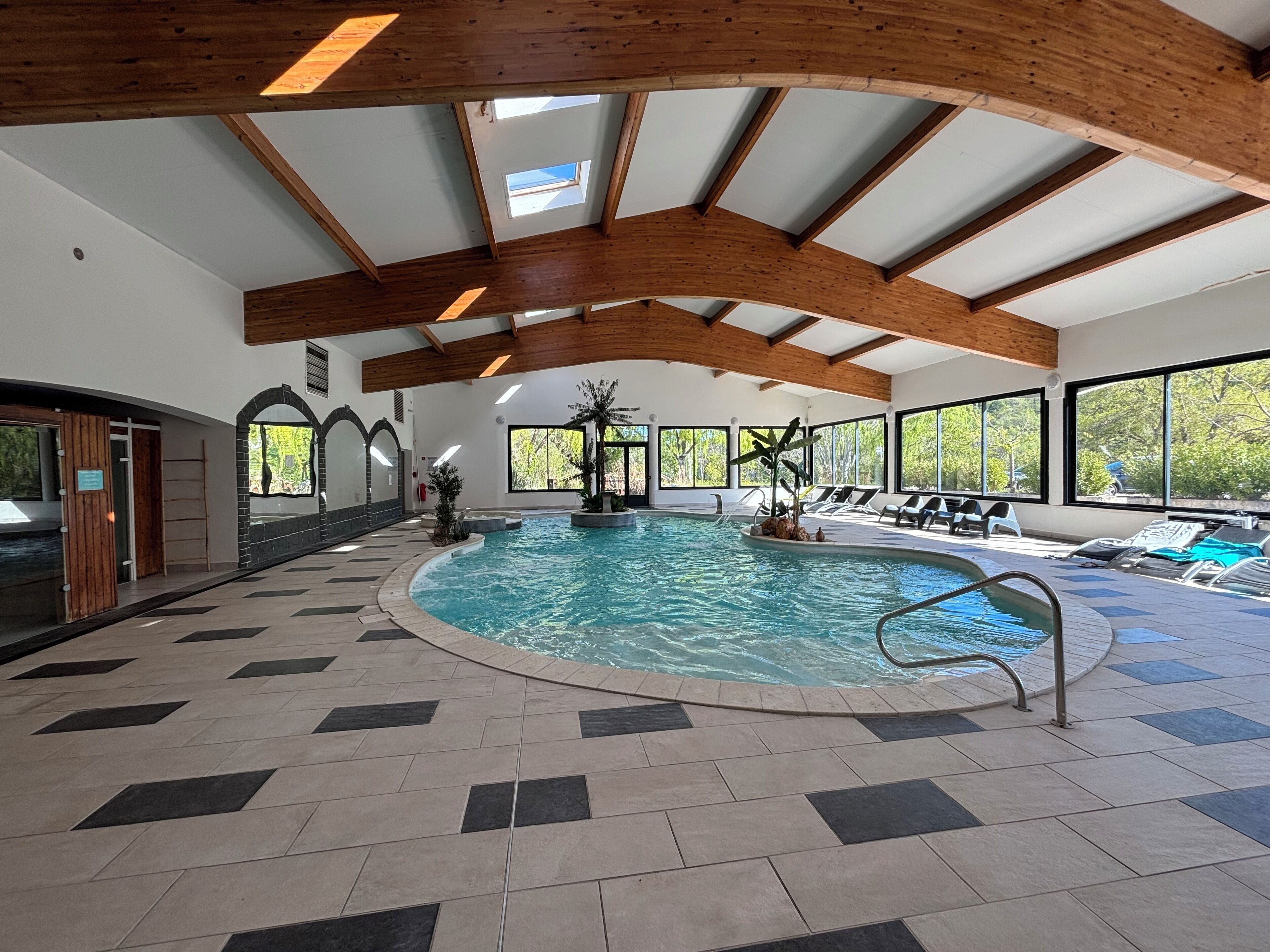 An indoor swimming pool with a tiled floor, wooden beams, and large windows.