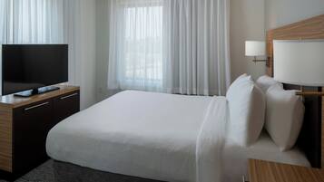 A hotel room with a large bed, a TV on a wooden desk, a bedside lamp, and a window with sheer curtains.