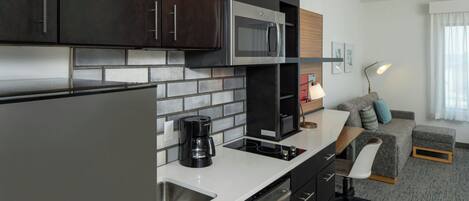 A modern kitchen with dark cabinets, a stainless steel refrigerator, a microwave, and a coffee maker. There is a small dining area with a table and a chair.
