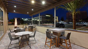 Un patio al aire libre con mesas y sillas, una pérgola en lo alto y vista a la calle y edificios al fondo.