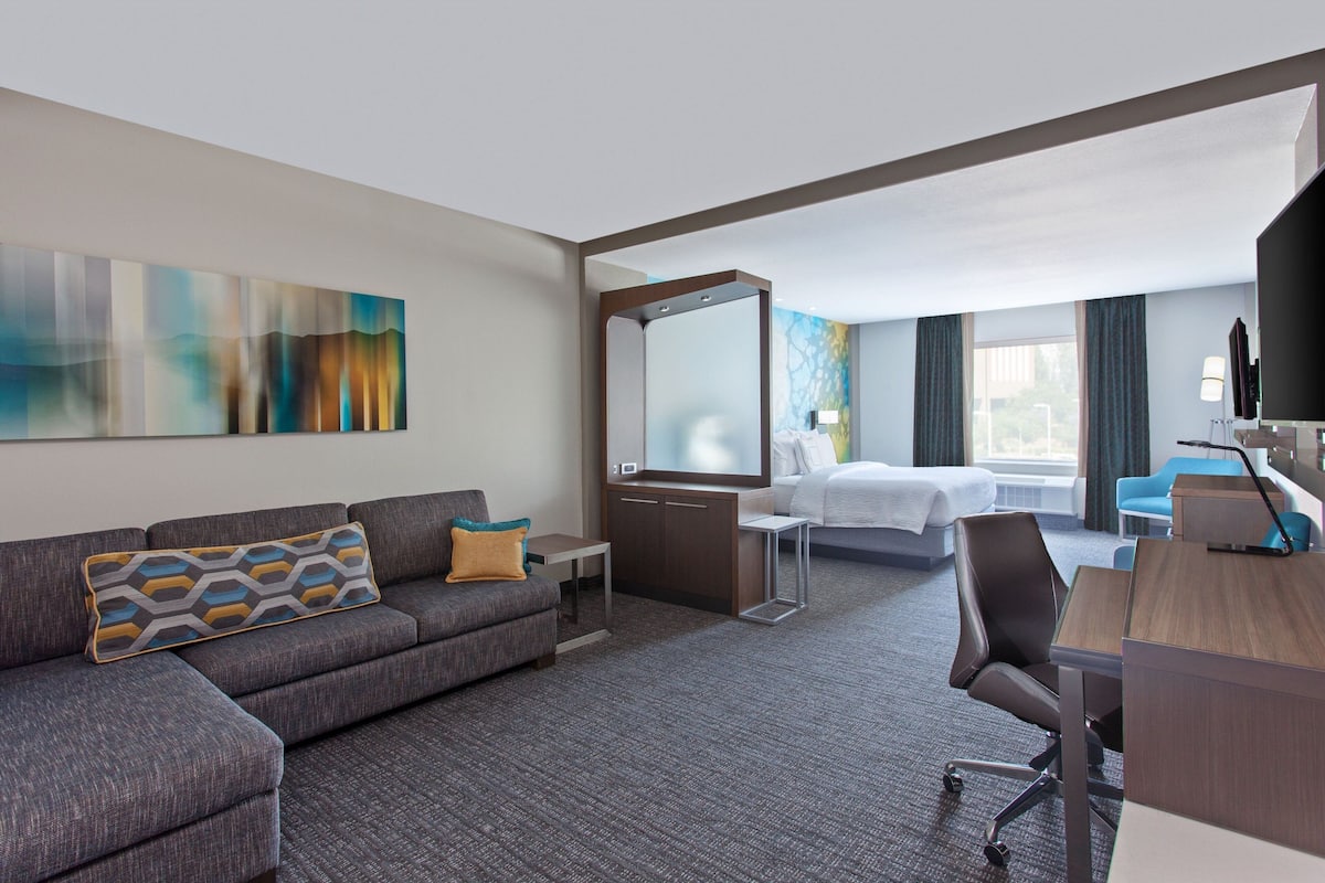 A modern hotel room with a sofa, a desk, a bed, a large mirror, and a painting on the wall.