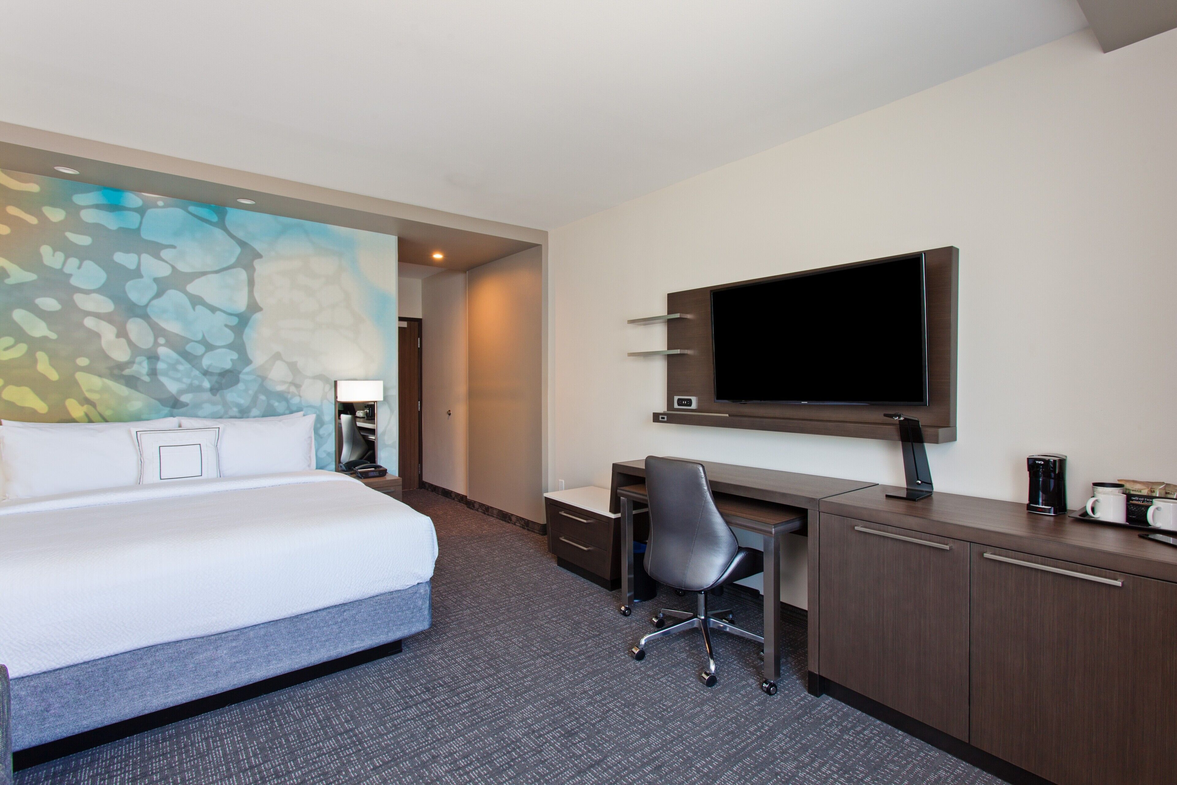 A hotel room with a bed, desk, chair, TV, and a painting on the wall.