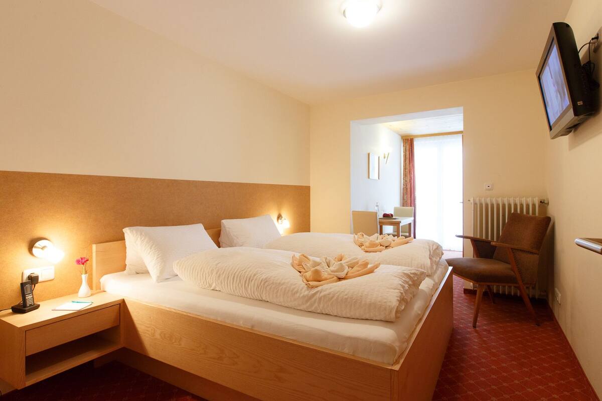 Een hotelkamer met een tweepersoonsbed, nachtkastjes met lampen, een stoel, een aan de muur bevestigde televisie en een radiator.