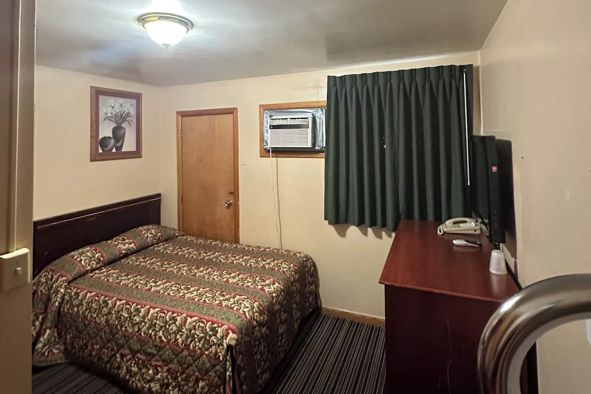 Quarto de hotel com cama, escrivaninha, cadeira, telefone, ar condicionado, janela com cortinas e luminária no teto.
