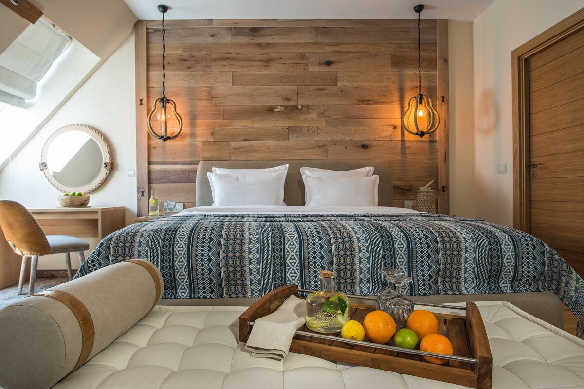 Ein Schlafzimmer mit einem Holz-Kopfteil, einem Bett mit gemusterten Betttextilien, einem Nachttisch mit Spiegel und einer Ablage mit Früchten und einer Flasche.