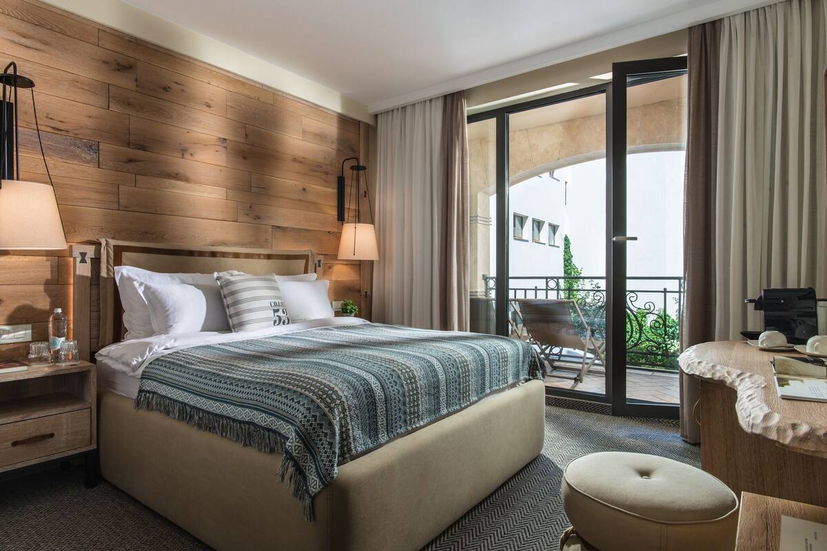 Ein Hotelzimmer mit einem hölzernen Kopfteil, einem großen Bett mit gemustertem Überwurf, einem Nachttisch mit Lampe, einem Schreibtisch mit Stuhl und einem Balkon mit Blick.