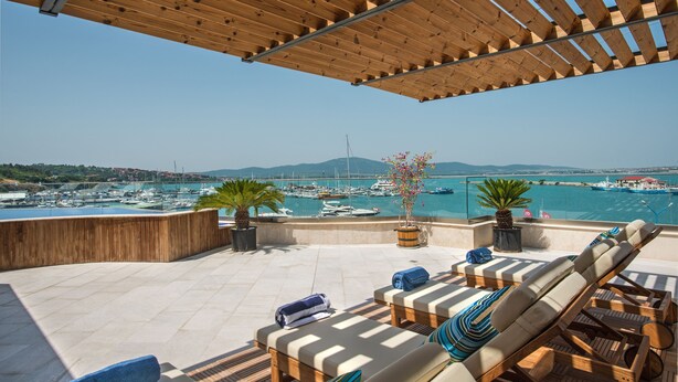 Une terrasse dotée de mobilier en bois, de plantes en pot et offrant une vue sur un port de plaisance avec des bateaux.