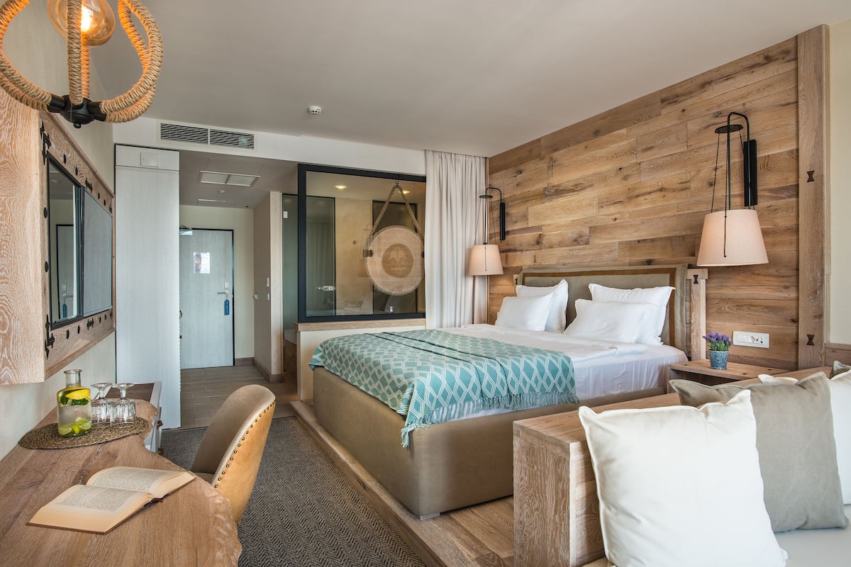 Ein modernes Hotelzimmer mit einer Wand in Holzoptik, einem großen Bett mit grünem Muster, einem Nachttisch mit Lampe sowie einer Sitzecke mit Sessel und kleinem Tisch.