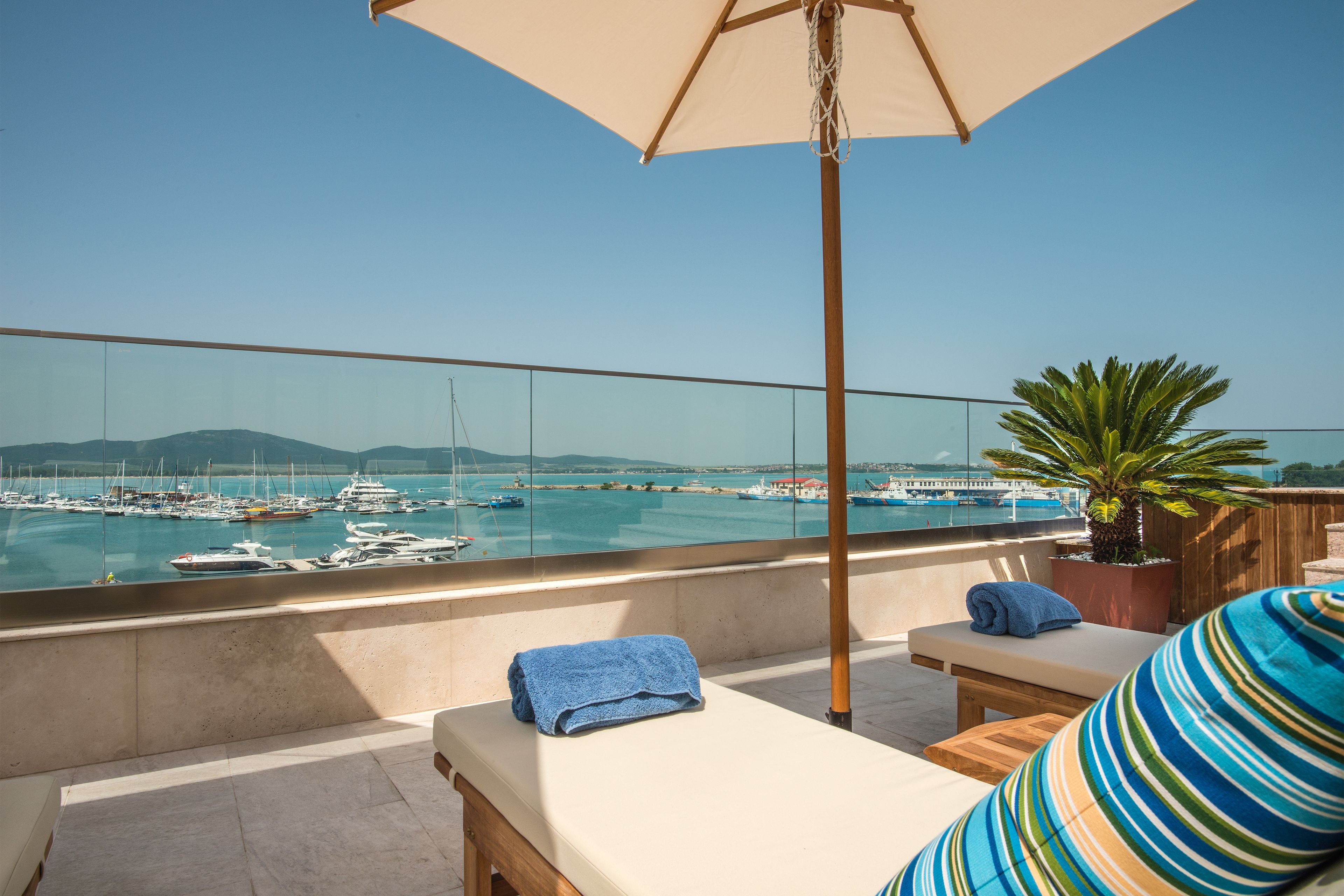 A balcony with a view of a marina, a potted plant, and lounge chairs with towels.