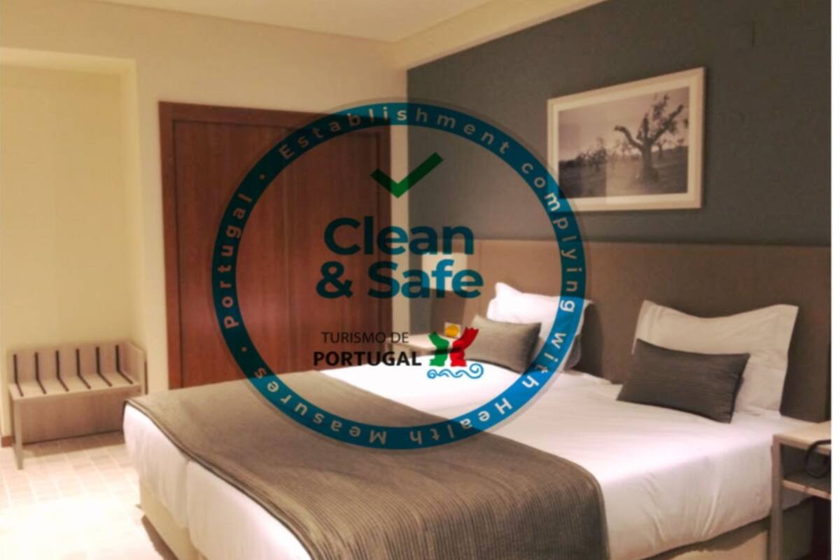A hotel room with a bed, a nightstand, and a chair. There is a logo with the text "Clean & Safe" and "Turismo de Portugal" on the wall.