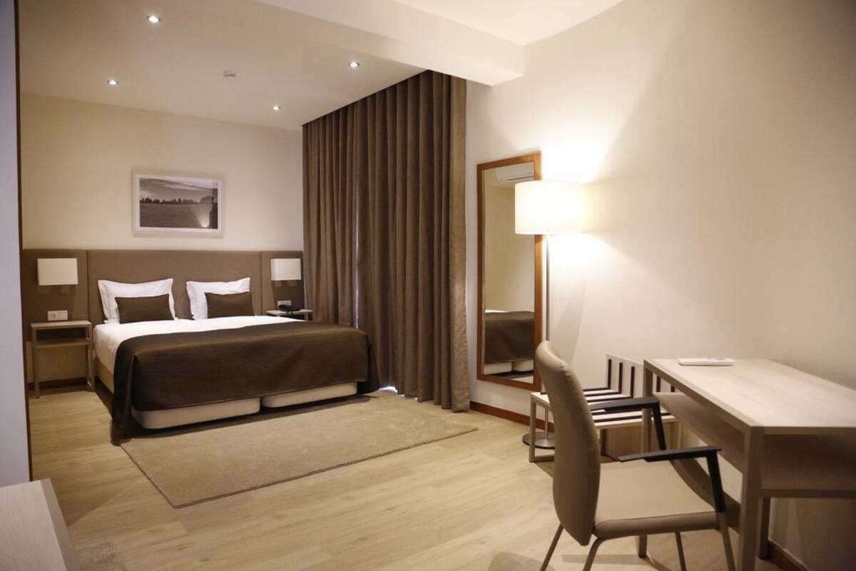A hotel room with a bed, bedside tables, a desk, a chair, a mirror, and a picture on the wall.